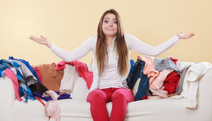 woman sitting on a couch shrugging while surrounded by extra clothes